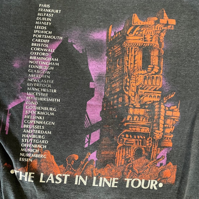 May include: A black t-shirt featuring a graphic of a futuristic city and the text "The Last in Line Tour." The graphic is in shades of orange and purple. The text is in white.