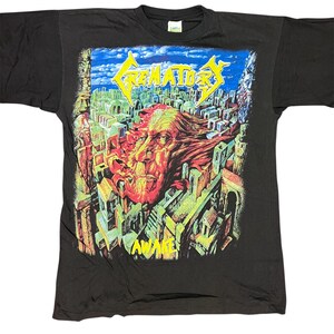 May include: A black short-sleeve t-shirt featuring a colorful graphic print. The design displays the text CREMATORY in yellow above a surreal illustration of a large face emerging from a city landscape with the word AWAKE printed in yellow below.