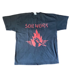 May include: Black t-shirt with the band name "SOILWORK" in red block letters. Below the text is a red graphic of a fist holding a knife, surrounded by flames and wings.