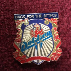 May include: A blue and gold metal pin with a red and white phoenix design. The pin features the text "Dokken" and "Back for the Attack".
