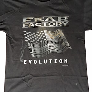 Puede incluir: Camiseta negra con un gráfico plateado de una bandera estadounidense ondeando con las palabras "FEAR FACTORY" y "REVOLUTION" impresas en ella.