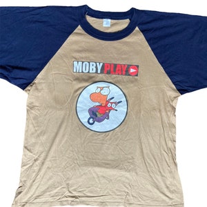 May include: A tan and navy blue baseball tee shirt with a graphic of a cartoon character riding a scooter. The text "MOBY PLAY" is printed in red and black on the shirt.