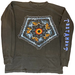 May include: Black long-sleeve t-shirt with a graphic print of a metal design with a black circle in the center. The text "TESTAMENT" is printed on the right sleeve in blue.