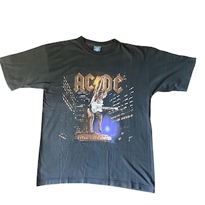 Puede incluir: Camiseta gris oscuro con la banda AC/DC. La camiseta tiene un gráfico de un guitarrista tocando frente a un paisaje urbano. El logotipo de la banda está en letras doradas encima de la imagen.