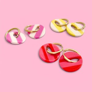 May include: A collection of colorful, striped earrings with gold hoops. The earrings are in shades of pink, yellow, and red, with white accents. The earrings are arranged on a solid pink background.