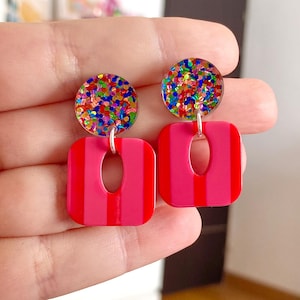Peut inclure: Une paire de boucles d'oreilles vives et colorées. Chaque boucle d'oreille présente un dessus rond et scintillant et un bas carré et rayé avec une découpe ovale. Les boucles d'oreilles sont suspendues à un crochet argenté. La palette de couleurs comprend des nuances de rose, de rouge et un arc-en-ciel de paillettes.