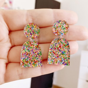May include: A pair of rainbow glitter earrings with a round top and a slightly arched bottom. The earrings are made of clear resin and are filled with colorful glitter.