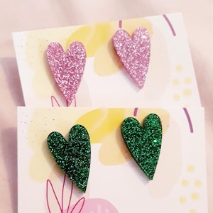 May include: Two pairs of glitter heart earrings. One pair is pink glitter and the other pair is green glitter. The earrings are displayed on a white card with a pink and yellow abstract design. The card also has the text "LA BOUTIQUE DE JUL" printed on it.