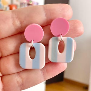 Puede incluir: Pendientes cuadrados a rayas rosas y azules con un círculo rosa en la parte superior. Los pendientes tienen un gancho plateado y un recorte en el centro del cuadrado. Los pendientes tienen una combinación de colores pastel.