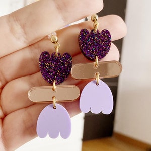 Puede incluir: Pendientes colgantes de color morado y lavanda con un corazón de purpurina dorada, un rectángulo de espejo y una forma de flor lavanda.
