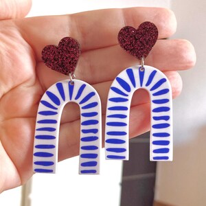 Peut inclure: Une paire de boucles d'oreilles fantaisie avec un cœur rouge pailleté et un motif d'arche blanche à rayures bleues. Les boucles d'oreilles sont tenues par une main, mettant en valeur le design. Un accessoire audacieux et coloré.