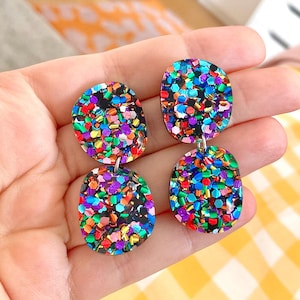 Peut inclure: Une paire de boucles d'oreilles scintillantes et colorées. Chaque boucle d'oreille présente une forme circulaire supérieure et une forme ovale inférieure, toutes deux remplies d'une mosaïque de paillettes aux couleurs de l'arc-en-ciel. Les boucles d'oreilles sont suspendues à un crochet argenté.