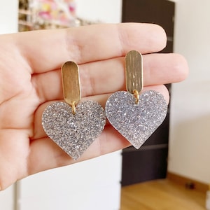Peut inclure: Une paire de boucles d'oreilles en forme de cœur avec des paillettes argentées et une barre doré sur le dessus