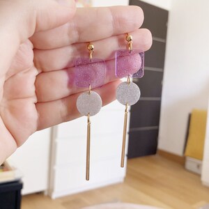 May include: A pair of dangle earrings with a square-shaped purple glitter charm and a round silver glitter charm, each with a gold bar hanging below.