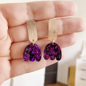 Peut inclure: Une paire de boucles d'oreilles pendantes dorées et violettes. Les boucles d'oreilles présentent une barre dorée avec un charme en forme de tulipe violet iridescent.
