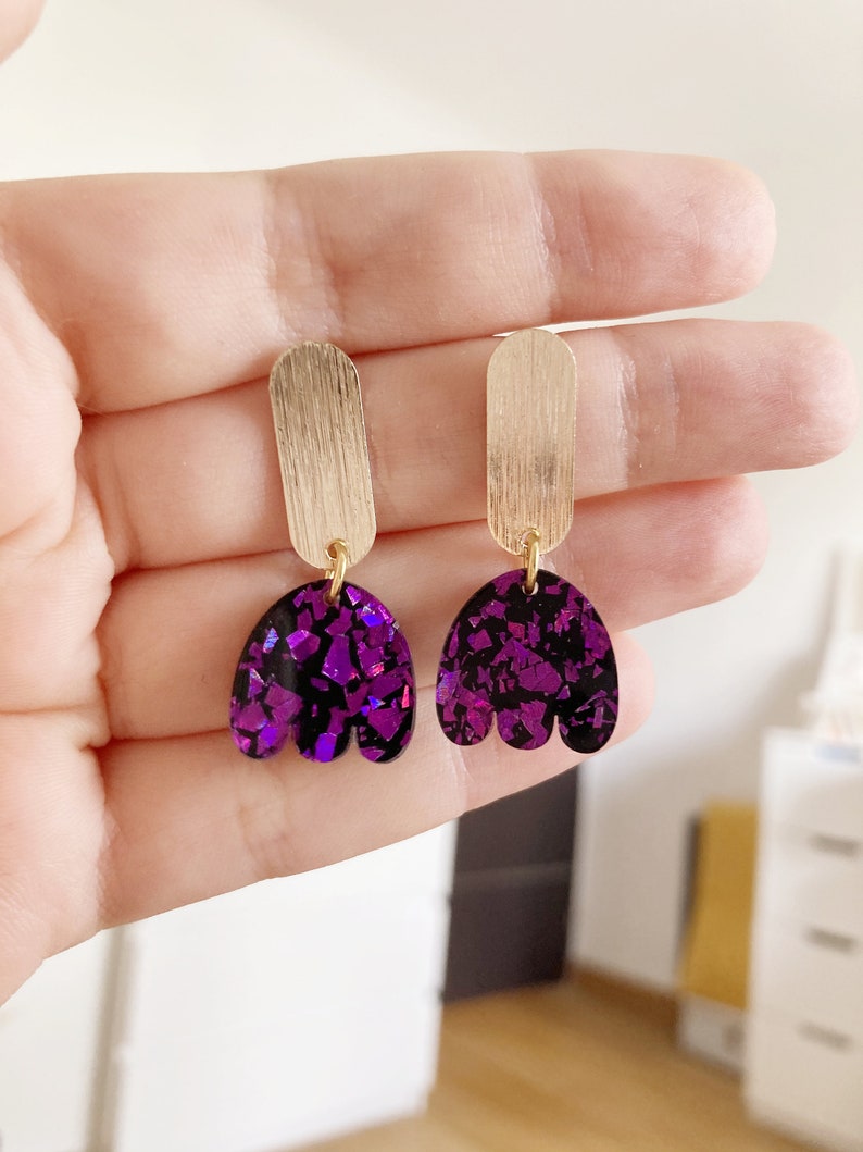 Peut inclure: Une paire de boucles d'oreilles pendantes dorées et violettes. Les boucles d'oreilles présentent une barre dorée avec un charme en forme de tulipe violet iridescent.