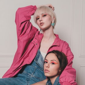 May include: Two women wearing denim overalls and a pink jacket. One woman has blonde hair and is wearing blue heart-shaped earrings. The other woman has brown hair and is wearing a silver ring on her finger.