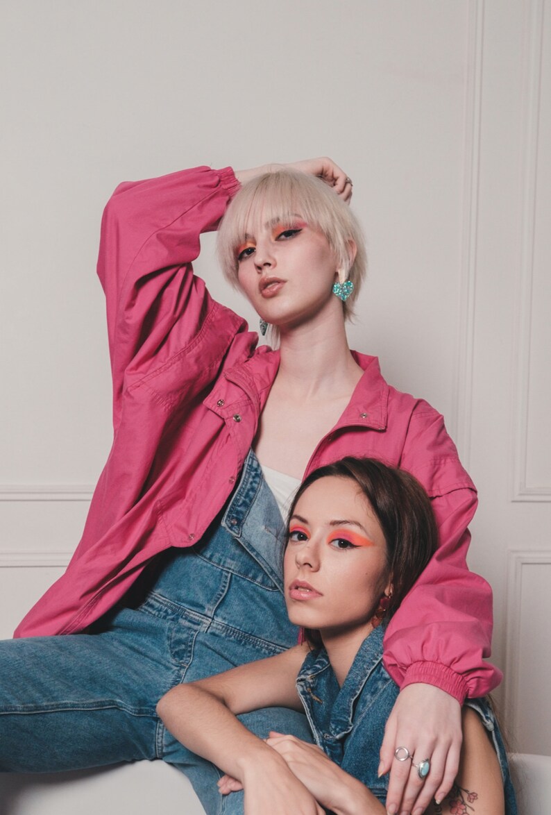 May include: Two women wearing denim overalls and a pink jacket. One woman has blonde hair and is wearing blue heart-shaped earrings. The other woman has brown hair and is wearing a silver ring on her finger.