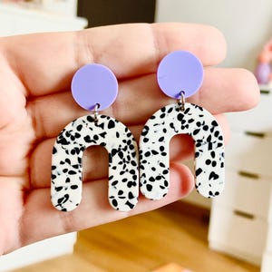 May include: A pair of dangle earrings with a white and black speckled arch shape and a lavender circle at the top.