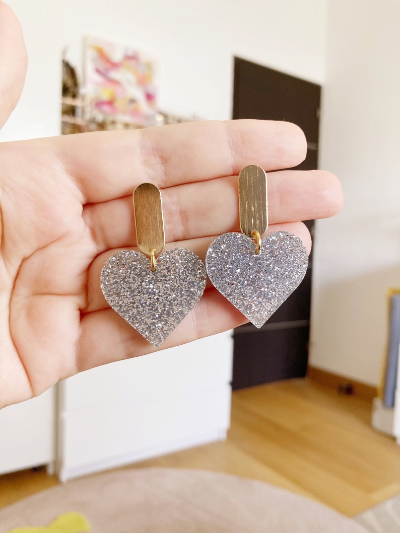 Peut inclure: Une paire de boucles d'oreilles en forme de cœur avec des paillettes argentées et une barre doré sur le dessus