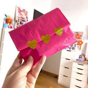 May include: A rectangular envelope made of bright pink paper, featuring a flap secured by three gold-colored heart-shaped stickers. The center heart sticker has a distinct iridescent, holographic texture, while the other two are solid gold.