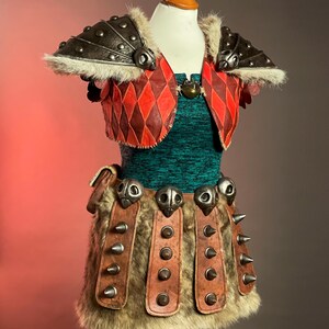 May include: A Viking-themed costume on a mannequin. The outfit includes a green textured top, a red and brown diamond-patterned vest, and a skirt with fur and leather details. The shoulders have metal armour with fur trim.