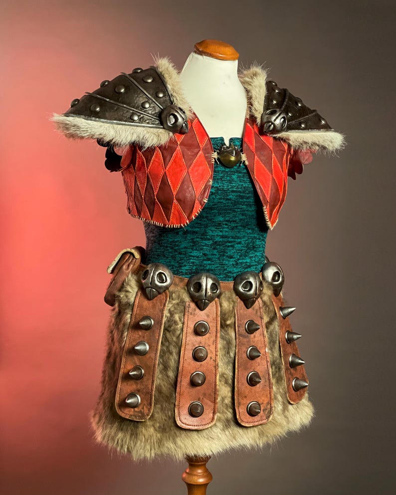 May include: A Viking-themed costume on a mannequin. The outfit includes a green textured top, a red and brown diamond-patterned vest, and a skirt with fur and leather details. The shoulders have metal armour with fur trim.