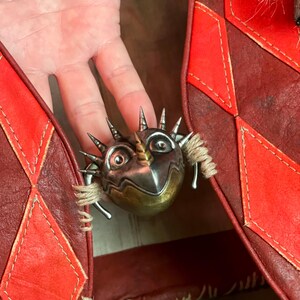 May include: A silver and gold-toned metal brooch shaped like a fantastical creature with spikes and a horn. The brooch is set against a red leather background with diamond-shaped accents.