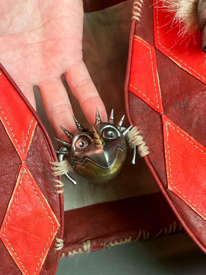 May include: A silver and gold-toned metal brooch shaped like a fantastical creature with spikes and a horn. The brooch is set against a red leather background with diamond-shaped accents.