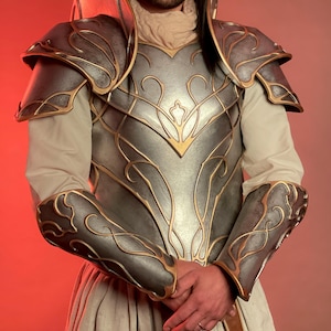 May include: A silver and gold fantasy armor set. The armor features intricate gold detailing on the chest, shoulders, and arm guards. The set includes a breastplate, shoulder pauldrons, and arm guards, all with a metallic finish.
