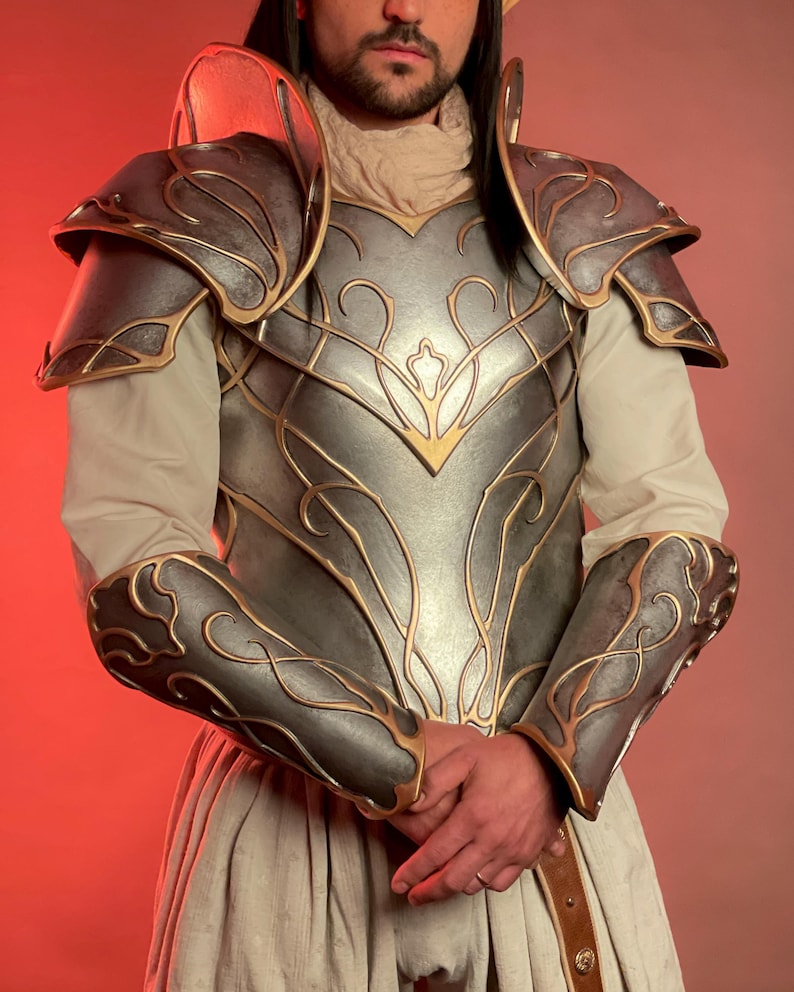May include: A silver and gold fantasy armor set. The armor features intricate gold detailing on the chest, shoulders, and arm guards. The set includes a breastplate, shoulder pauldrons, and arm guards, all with a metallic finish.