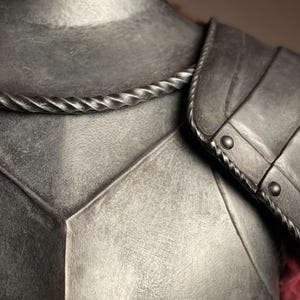 May include: Close-up of a metallic gray medieval armor chest piece. The armor features a textured surface, a decorative rope-like trim around the neck, and a shoulder guard with rivets. The design suggests historical or fantasy themes.