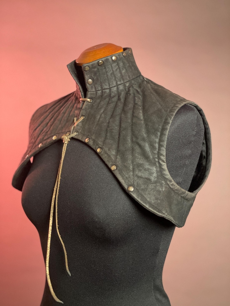 以下が含まれることがあります： 黒い革製のショルダーピース。ハイカラー、キルティングデザイン、金属スタッド付き。 レースアップ開閉式で、黒いマネキンに展示されています。 全体的なテーマは中世またはファンタジーです。