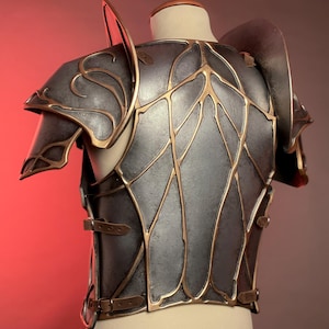 May include: A detailed, metallic gray and gold breastplate and shoulder armor. The armor features intricate gold leaf-like designs and is secured with leather straps and buckles. The shoulder pieces have curved edges and are mounted on a mannequin.