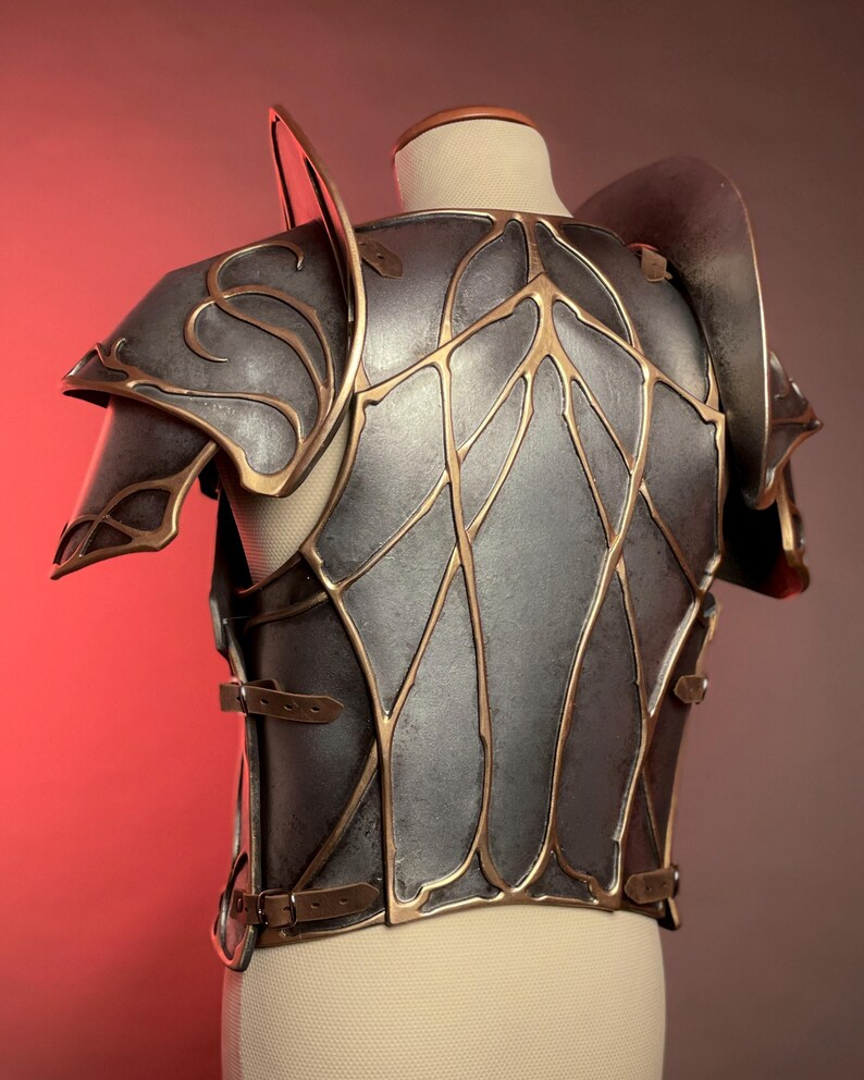 May include: A detailed, metallic gray and gold breastplate and shoulder armor. The armor features intricate gold leaf-like designs and is secured with leather straps and buckles. The shoulder pieces have curved edges and are mounted on a mannequin.
