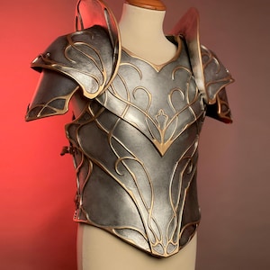 May include: A silver-colored breastplate and shoulder armor with gold-colored filigree and trim. The armor is displayed on a mannequin and has a detailed, ornate design. The shoulder pieces are large and curved, and the breastplate has a central point.