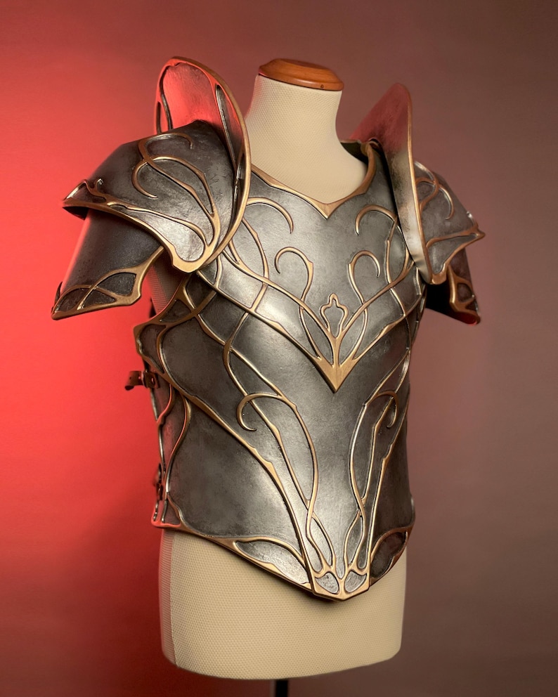May include: A silver-colored breastplate and shoulder armor with gold-colored filigree and trim. The armor is displayed on a mannequin and has a detailed, ornate design. The shoulder pieces are large and curved, and the breastplate has a central point.