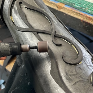 May include: Close-up of a craft project in progress. A rotary tool is being used on a dark gray, cylindrical object with black, decorative cutouts. The object appears to be part of a larger artistic creation.