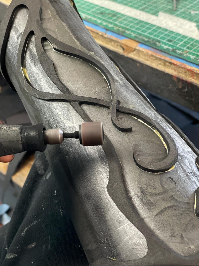 May include: Close-up of a craft project in progress. A rotary tool is being used on a dark gray, cylindrical object with black, decorative cutouts. The object appears to be part of a larger artistic creation.