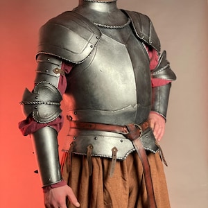 May include: A detailed, metallic gray medieval armor set. The armor includes a chest plate, shoulder guards, arm guards, and a neck piece. A brown leather belt and tan fabric are also visible. The armor is designed with rivets and decorative edges.