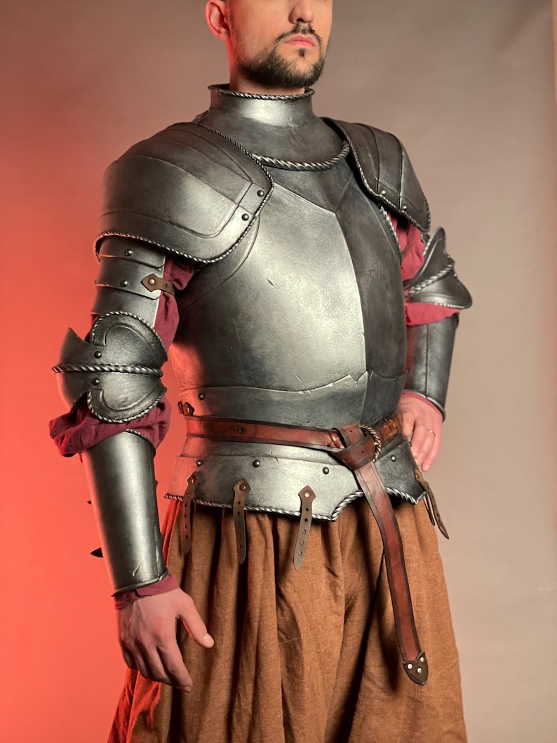 May include: A detailed, metallic gray medieval armor set. The armor includes a chest plate, shoulder guards, arm guards, and a neck piece. A brown leather belt and tan fabric are also visible. The armor is designed with rivets and decorative edges.
