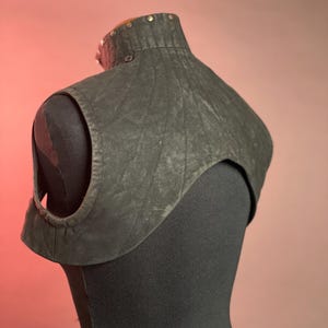 以下が含まれることがあります： 高いスタッズ付きのネックラインを備えた、ダークグレーの質感のあるレザーカラー。カラーは肩の部分にキルティングデザインがあり、小さなボタンで留めます。この衣服は黒いマネキンに展示されています。