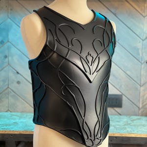 May include: Black chest armor with an intricate, raised floral design. The armor is displayed on a mannequin and has a V-neck. The design features curved lines and leaf-like shapes, creating a detailed and textured appearance.