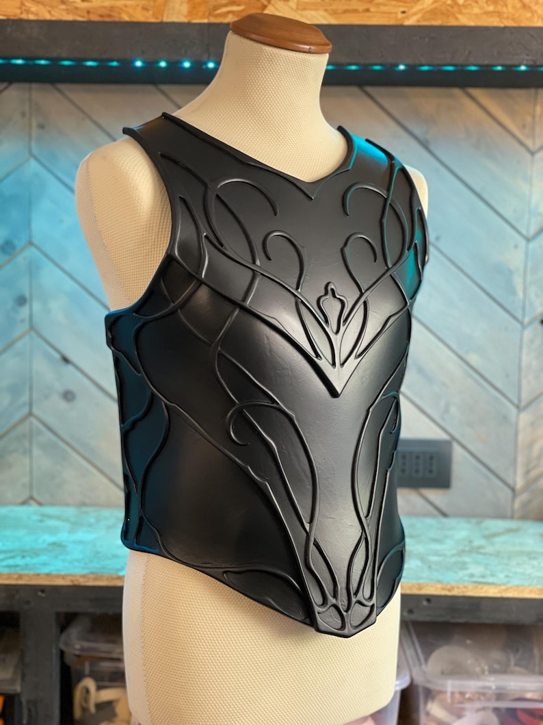 May include: Black chest armor with an intricate, raised floral design. The armor is displayed on a mannequin and has a V-neck. The design features curved lines and leaf-like shapes, creating a detailed and textured appearance.