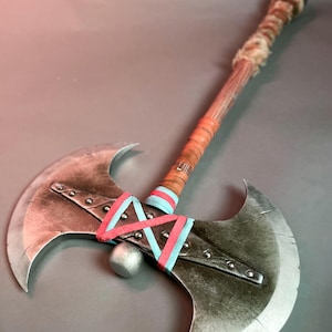 May include: A double-bladed axe with a wooden handle wrapped in leather and fur. The axe heads are silver with a distressed finish, and the handle is decorated with red and blue ribbons. A silver ball is between the axe heads.