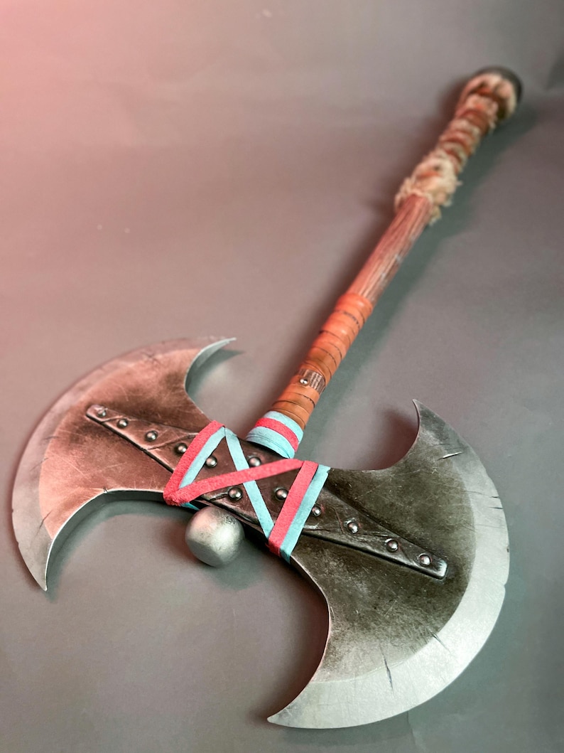 May include: A double-bladed axe with a wooden handle wrapped in leather and fur. The axe heads are silver with a distressed finish, and the handle is decorated with red and blue ribbons. A silver ball is between the axe heads.