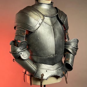 May include: A detailed, silver-colored suit of armor, displayed on a mannequin. The armor features articulated plates, rivets, and leather straps. The design includes shoulder guards, arm protection, and a chest plate, showcasing medieval craftsmanship.