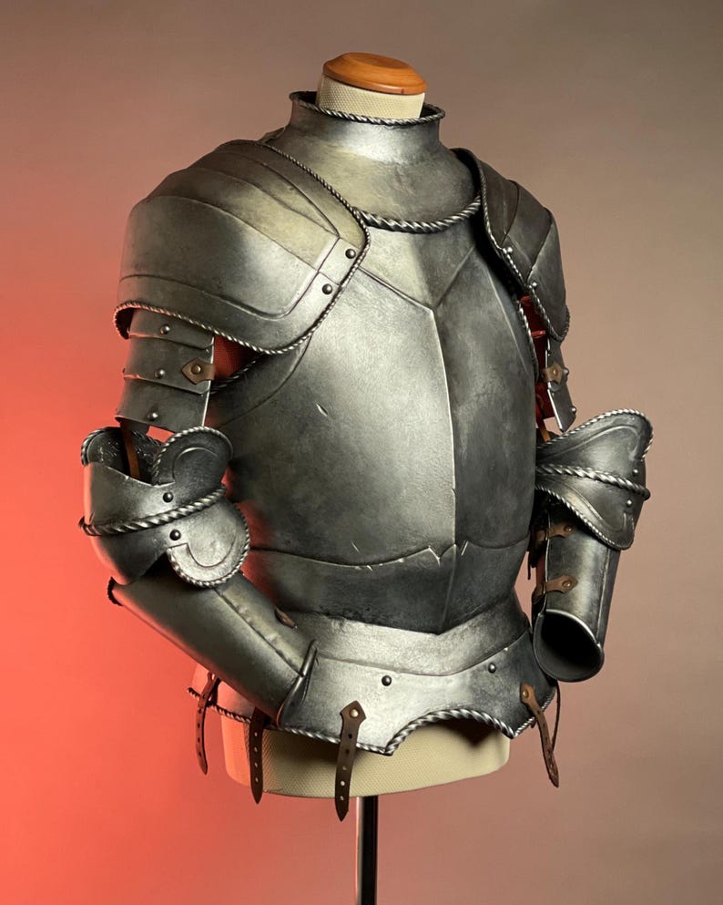 May include: A detailed, silver-colored suit of armor, displayed on a mannequin. The armor features articulated plates, rivets, and leather straps. The design includes shoulder guards, arm protection, and a chest plate, showcasing medieval craftsmanship.