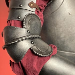 May include: Close-up of a medieval-style arm armor. The dark metal armor features articulated plates, decorative rope-like trim, and a leather strap. A burgundy fabric is visible beneath the armor plates, adding a touch of color.