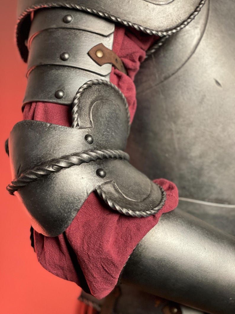 May include: Close-up of a medieval-style arm armor. The dark metal armor features articulated plates, decorative rope-like trim, and a leather strap. A burgundy fabric is visible beneath the armor plates, adding a touch of color.
